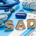 Medical tools and symbols representing Seasonal Affective Disorder (SAD) therapy. The word 'SAD' is spelled in wooden letters on a blue background, surrounded by a stethoscope, a model brain, a reflex hammer, and a strip of white pills. A mental health assessment form partially visible highlights clinical aspects of SAD diagnosis and treatment.