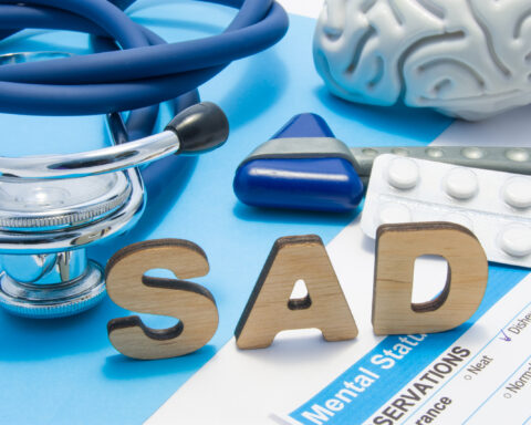 Medical tools and symbols representing Seasonal Affective Disorder (SAD) therapy. The word 'SAD' is spelled in wooden letters on a blue background, surrounded by a stethoscope, a model brain, a reflex hammer, and a strip of white pills. A mental health assessment form partially visible highlights clinical aspects of SAD diagnosis and treatment.