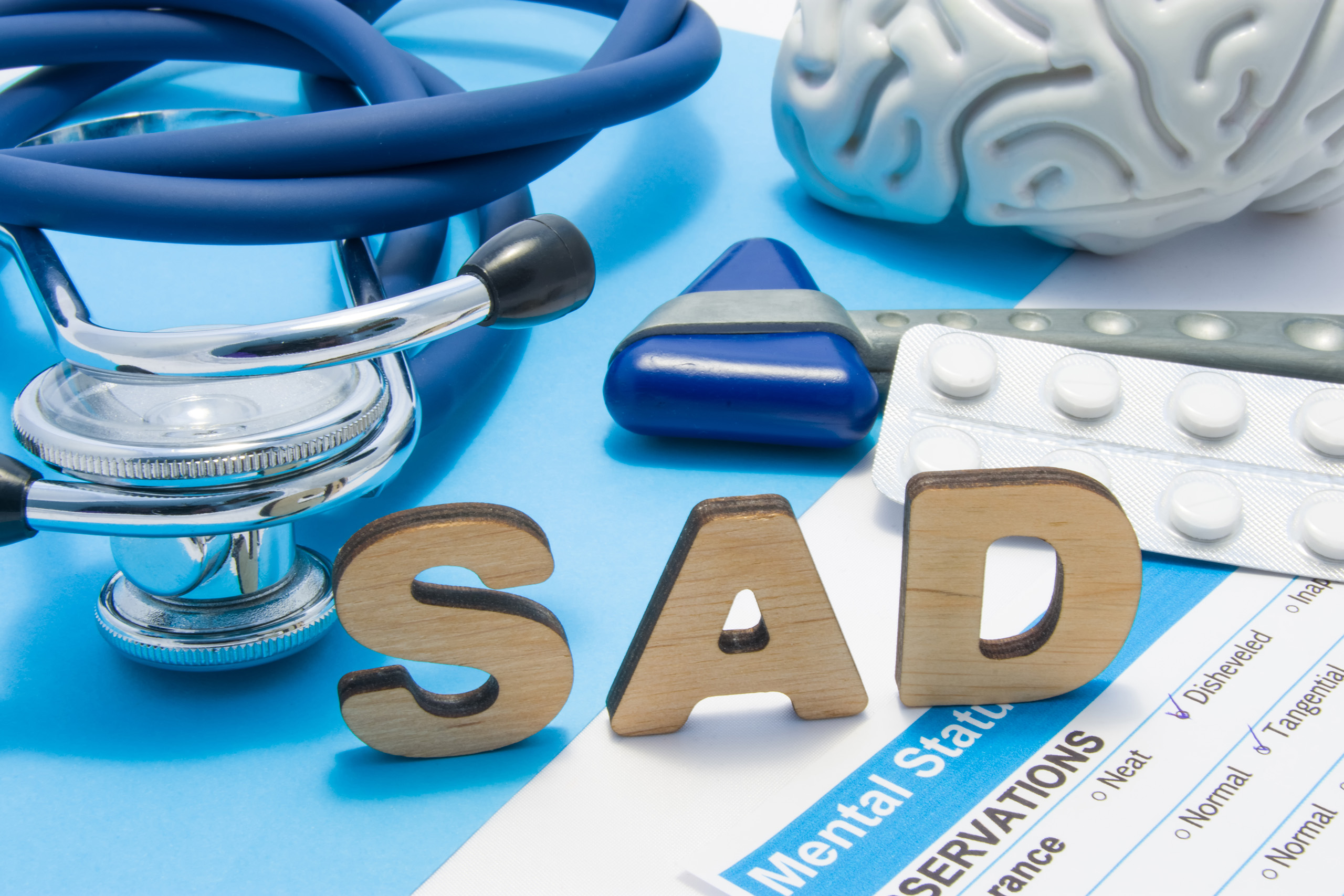 Medical tools and symbols representing Seasonal Affective Disorder (SAD) therapy. The word 'SAD' is spelled in wooden letters on a blue background, surrounded by a stethoscope, a model brain, a reflex hammer, and a strip of white pills. A mental health assessment form partially visible highlights clinical aspects of SAD diagnosis and treatment.