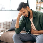 A man sits on a couch in casual clothes, holding his stomach with one hand while resting his head on the other in a gesture of discomfort. His facial expression suggests he is experiencing pain or distress, possibly due to gastrointestinal issues. The cozy, softly lit background of the living room contrasts with his obvious discomfort, indicating a health concern within a home setting.