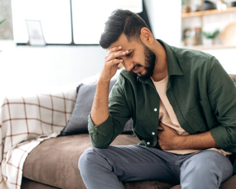 A man sits on a couch in casual clothes, holding his stomach with one hand while resting his head on the other in a gesture of discomfort. His facial expression suggests he is experiencing pain or distress, possibly due to gastrointestinal issues. The cozy, softly lit background of the living room contrasts with his obvious discomfort, indicating a health concern within a home setting.