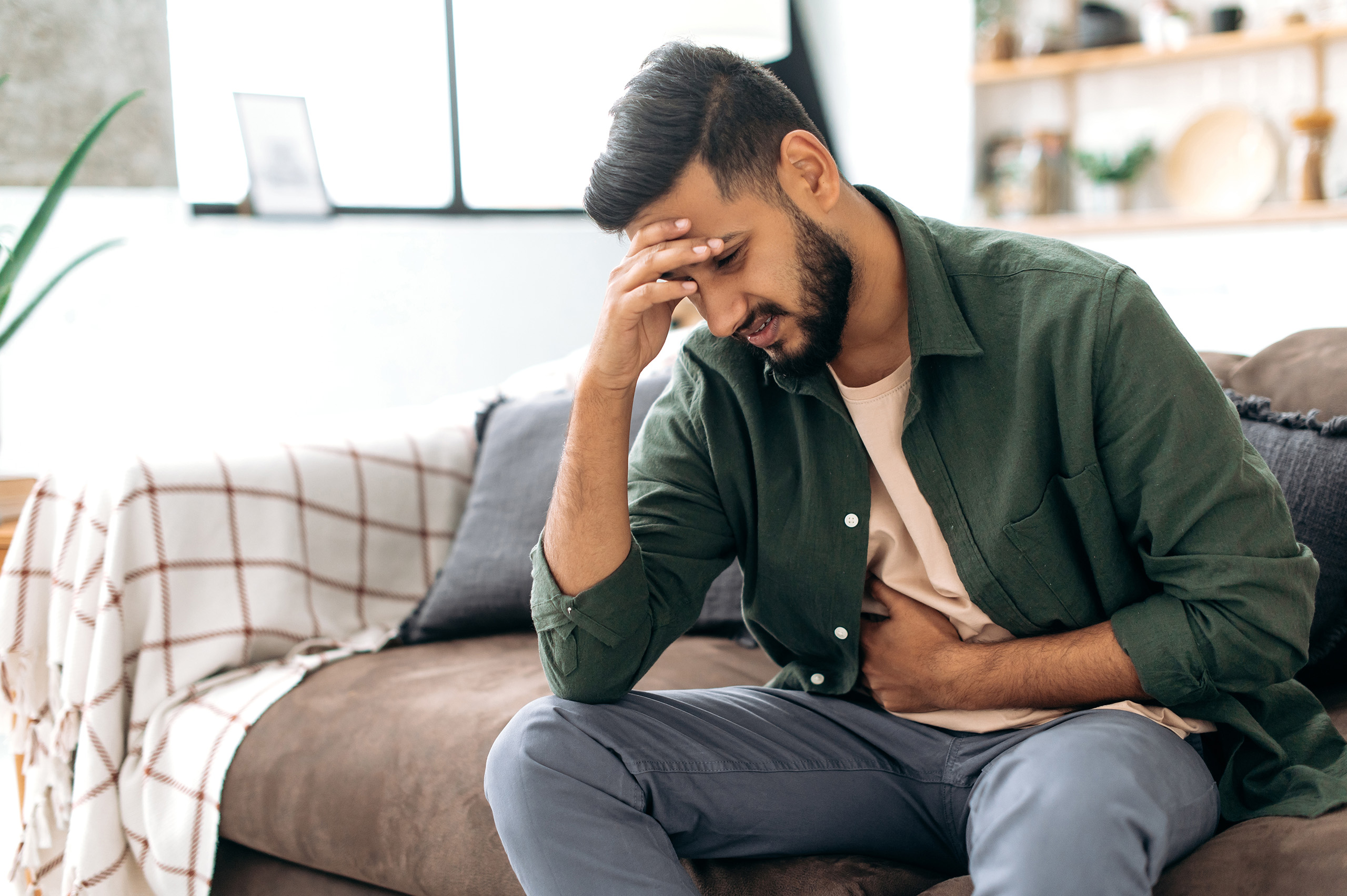 A man sits on a couch in casual clothes, holding his stomach with one hand while resting his head on the other in a gesture of discomfort. His facial expression suggests he is experiencing pain or distress, possibly due to gastrointestinal issues. The cozy, softly lit background of the living room contrasts with his obvious discomfort, indicating a health concern within a home setting.