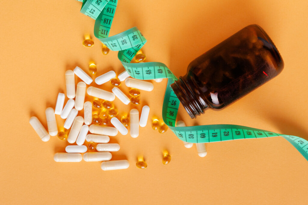 An amber-colored bottle of dietary supplements spills white capsules and golden softgels onto a light orange surface, with a green measuring tape draped around it. The image evokes themes of health, wellness, and weight management, symbolizing the scientific pursuit of mimicking caloric restriction through supplements to promote longevity and vitality.