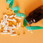 An amber-colored bottle of dietary supplements spills white capsules and golden softgels onto a light orange surface, with a green measuring tape draped around it. The image evokes themes of health, wellness, and weight management, symbolizing the scientific pursuit of mimicking caloric restriction through supplements to promote longevity and vitality.