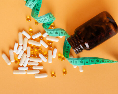 An amber-colored bottle of dietary supplements spills white capsules and golden softgels onto a light orange surface, with a green measuring tape draped around it. The image evokes themes of health, wellness, and weight management, symbolizing the scientific pursuit of mimicking caloric restriction through supplements to promote longevity and vitality.