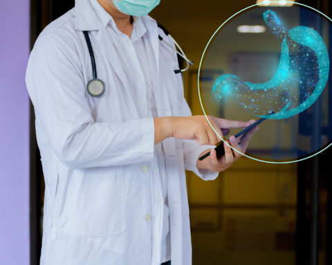 A doctor wearing a white coat and a surgical mask is holding a tablet and pointing at it. In the foreground, a digital hologram of a stomach, composed of a glowing blue wireframe, is superimposed. The image suggests the use of advanced medicine in gastroenterology or digestive health.