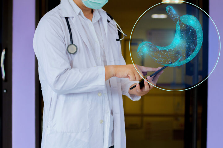 A doctor wearing a white coat and a surgical mask is holding a tablet and pointing at it. In the foreground, a digital hologram of a stomach, composed of a glowing blue wireframe, is superimposed. The image suggests the use of advanced medicine in gastroenterology or digestive health.