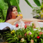 The image shows a serene outdoor scene where a woman is relaxing in a bath filled with water and rose petals. She is lounging in a white bathtub, surrounded by vibrant greenery and colorful flowers. The woman is holding a glass of detox juice and appears to be enjoying a peaceful moment. Nearby, there are spa-like items, such as towels and bottles of lotions, placed on a black tray. The setting is tropical, with large green leaves and a yellow bicycle visible in the background, giving the image a refreshing and tranquil spa ambiance.