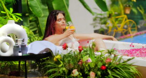 The image shows a serene outdoor scene where a woman is relaxing in a bath filled with water and rose petals. She is lounging in a white bathtub, surrounded by vibrant greenery and colorful flowers. The woman is holding a glass of detox juice and appears to be enjoying a peaceful moment. Nearby, there are spa-like items, such as towels and bottles of lotions, placed on a black tray. The setting is tropical, with large green leaves and a yellow bicycle visible in the background, giving the image a refreshing and tranquil spa ambiance.