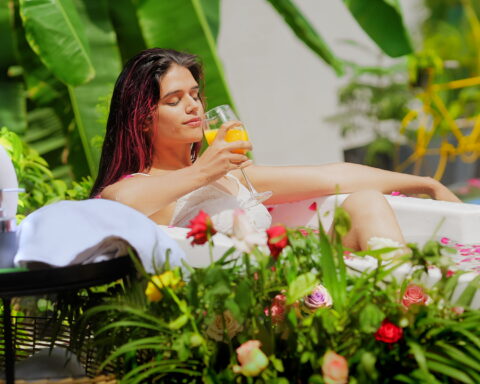 The image shows a serene outdoor scene where a woman is relaxing in a bath filled with water and rose petals. She is lounging in a white bathtub, surrounded by vibrant greenery and colorful flowers. The woman is holding a glass of detox juice and appears to be enjoying a peaceful moment. Nearby, there are spa-like items, such as towels and bottles of lotions, placed on a black tray. The setting is tropical, with large green leaves and a yellow bicycle visible in the background, giving the image a refreshing and tranquil spa ambiance.