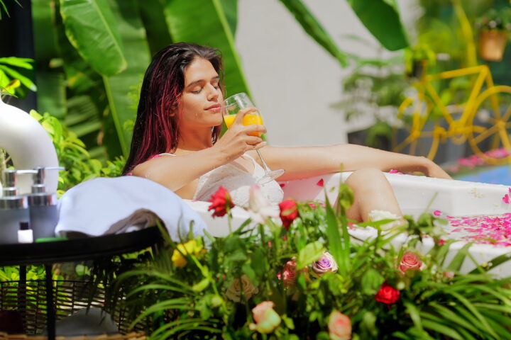 The image shows a serene outdoor scene where a woman is relaxing in a bath filled with water and rose petals. She is lounging in a white bathtub, surrounded by vibrant greenery and colorful flowers. The woman is holding a glass of detox juice and appears to be enjoying a peaceful moment. Nearby, there are spa-like items, such as towels and bottles of lotions, placed on a black tray. The setting is tropical, with large green leaves and a yellow bicycle visible in the background, giving the image a refreshing and tranquil spa ambiance.