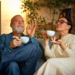 An older couple sitting on a comfortable couch, enjoying cups of tea or coffee, sharing a lighthearted and relaxed conversation. The man, wearing a denim jacket, smiles and gestures in delight, while the woman in glasses and a cream outfit engages animatedly. The warm and cozy indoor setting, with soft lighting and greenery in the background, evokes a sense of serenity and connection.