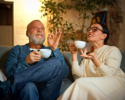 An older couple sitting on a comfortable couch, enjoying cups of tea or coffee, sharing a lighthearted and relaxed conversation. The man, wearing a denim jacket, smiles and gestures in delight, while the woman in glasses and a cream outfit engages animatedly. The warm and cozy indoor setting, with soft lighting and greenery in the background, evokes a sense of serenity and connection.