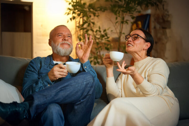 An older couple sitting on a comfortable couch, enjoying cups of tea or coffee, sharing a lighthearted and relaxed conversation. The man, wearing a denim jacket, smiles and gestures in delight, while the woman in glasses and a cream outfit engages animatedly. The warm and cozy indoor setting, with soft lighting and greenery in the background, evokes a sense of serenity and connection.