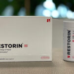 A promotional image showcasing the RESTORIN® product packaging and bottle. The white box with bold black text "RESTORIN SE" and the accompanying bottle labeled "Cellular Energy & Repair Aging Optimization" are placed on a neutral surface, with a blurred natural background that emphasizes innovation and health. The sleek, minimalist design of the packaging highlights RESTORIN®'s focus on advanced scientific research targeting the root causes of aging, including mTOR modulation, senolytics, and sirtuin activation.