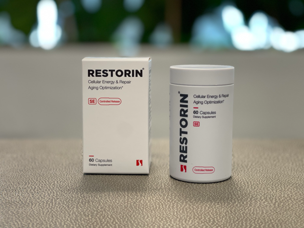 RESTORIN product display showcasing its premium design and packaging. The image features a white rectangular box labeled 'RESTORIN - Cellular Energy & Repair, Aging Optimization,' alongside a cylindrical bottle with matching minimalist aesthetics. Both are positioned on a sleek surface, with a blurred natural background emphasizing the clean and professional presentation. RESTORIN, developed by Seragon Biosciences, is described as a cutting-edge nutraceutical targeting healthy aging through advanced technologies like mTOR inhibition, autophagy activation, and senolytics.