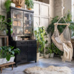 Cozy indoor space with lush houseplants including monstera, ferns, and snake plants. Features a hammock, rustic wooden furniture, and natural light from large windows.