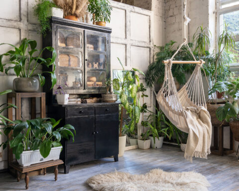 Cozy indoor space with lush houseplants including monstera, ferns, and snake plants. Features a hammock, rustic wooden furniture, and natural light from large windows.