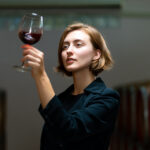 Young woman evaluating a glass of red wine in a winery, surrounded by wooden wine barrels, symbolizing the exploration of red wine's health benefits and its role in longevity.