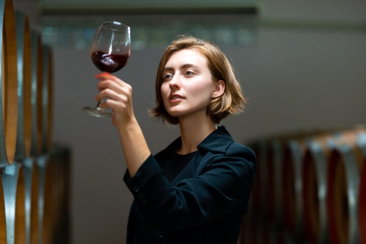 Young woman evaluating a glass of red wine in a winery, surrounded by wooden wine barrels, symbolizing the exploration of red wine's health benefits and its role in longevity.