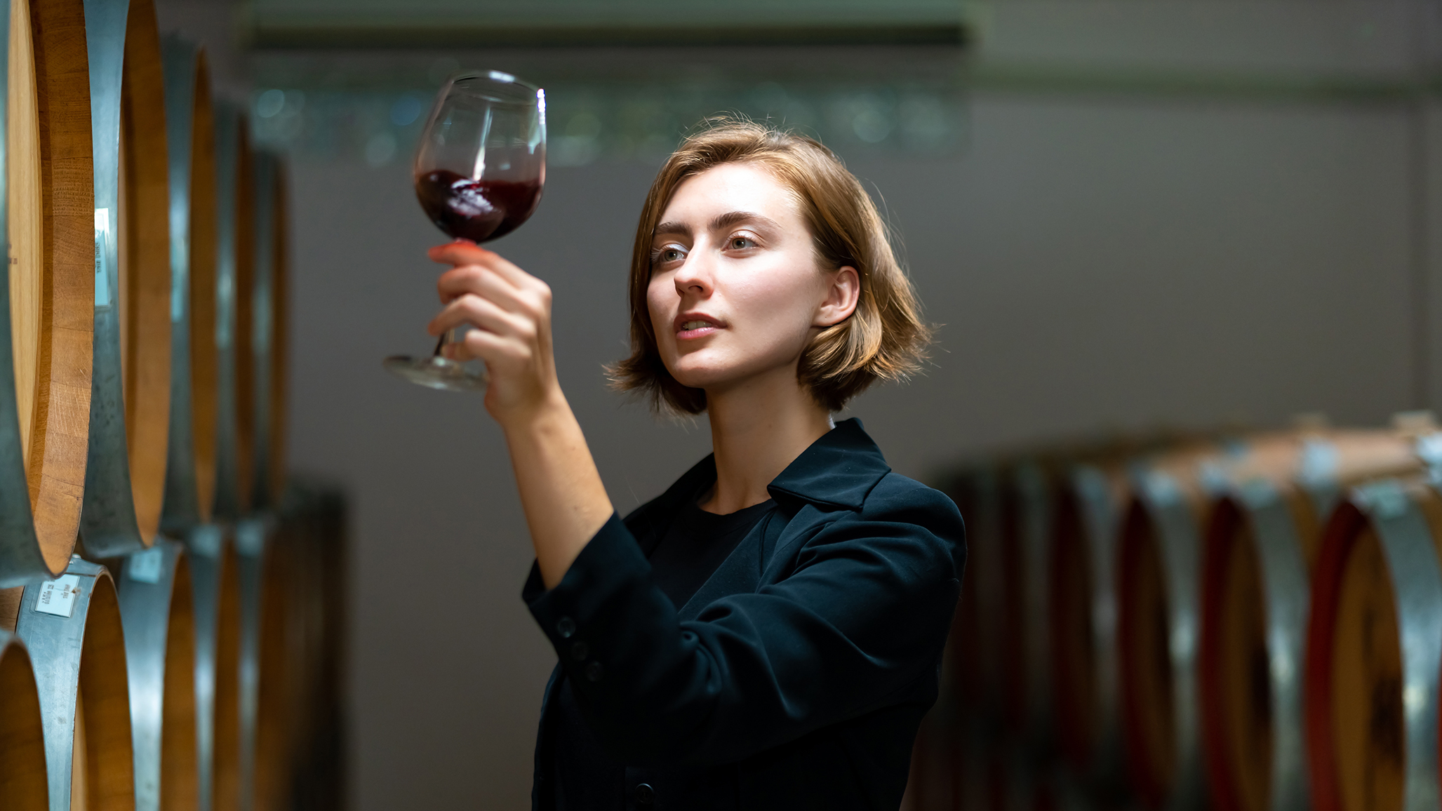 Young woman evaluating a glass of red wine in a winery, surrounded by wooden wine barrels, symbolizing the exploration of red wine's health benefits and its role in longevity.