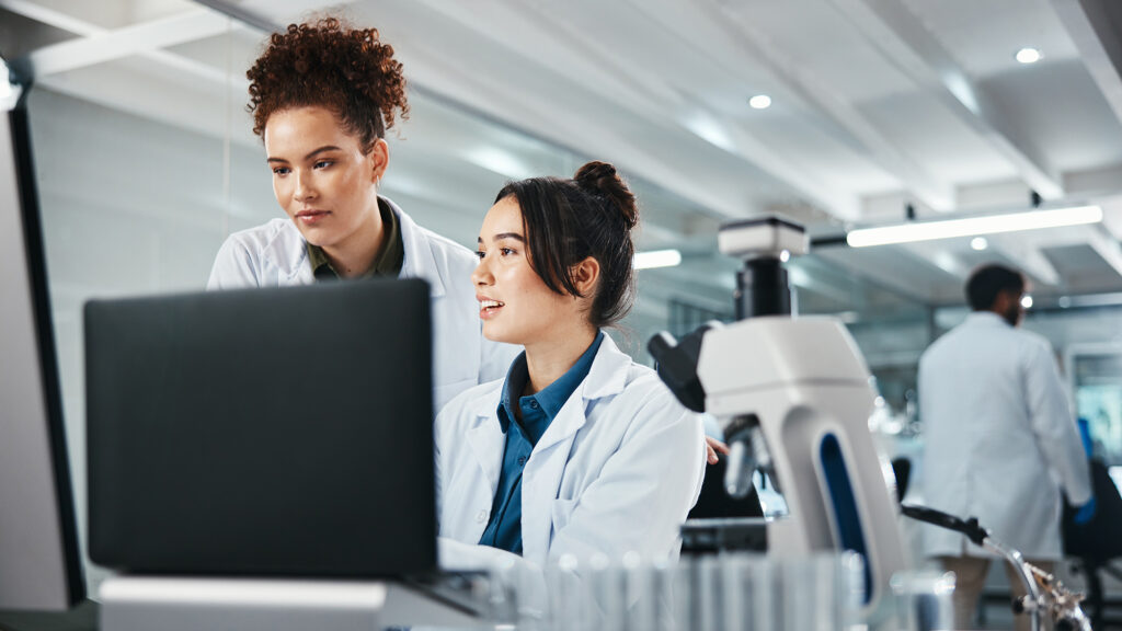 Two female scientists in lab coats analyze data on a computer in a modern laboratory, with a microscope and test tubes in the foreground. A male scientist works in the background.