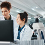 Two female scientists in lab coats analyze data on a computer in a modern laboratory, with a microscope and test tubes in the foreground. A male scientist works in the background.