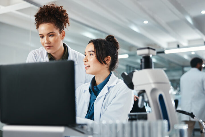 Two female scientists in lab coats analyze data on a computer in a modern laboratory, with a microscope and test tubes in the foreground. A male scientist works in the background.