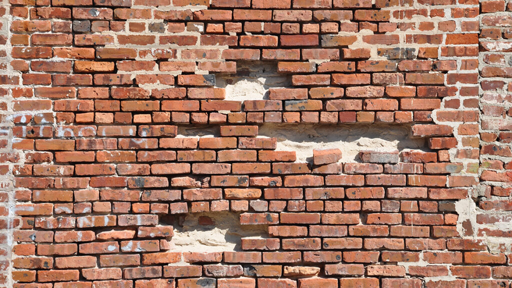 A weathered red brick wall with multiple missing and crumbling bricks, revealing gaps and structural weaknesses. This visually symbolizes genomic instability—how accumulating DNA damage over time weakens cellular function, leading to aging and disease. The deteriorating wall represents the gradual breakdown of the body's genetic integrity, akin to mutations and repair failures that drive the aging process.