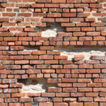 A weathered red brick wall with multiple missing and crumbling bricks, revealing gaps and structural weaknesses. This visually symbolizes genomic instability—how accumulating DNA damage over time weakens cellular function, leading to aging and disease. The deteriorating wall represents the gradual breakdown of the body's genetic integrity, akin to mutations and repair failures that drive the aging process.