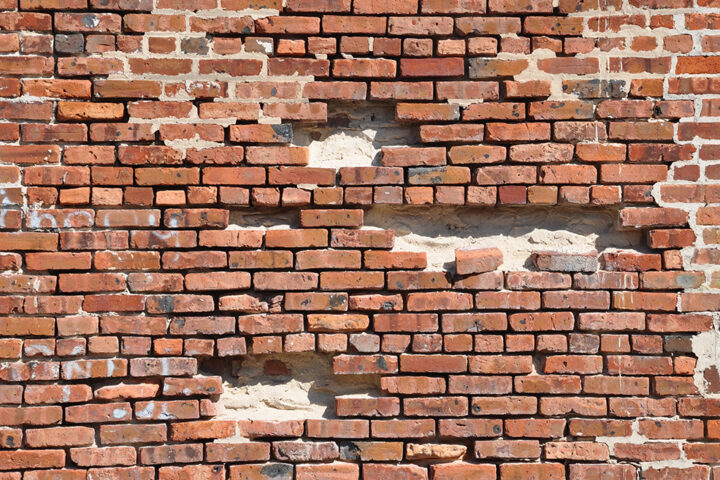 A weathered red brick wall with multiple missing and crumbling bricks, revealing gaps and structural weaknesses. This visually symbolizes genomic instability—how accumulating DNA damage over time weakens cellular function, leading to aging and disease. The deteriorating wall represents the gradual breakdown of the body's genetic integrity, akin to mutations and repair failures that drive the aging process.