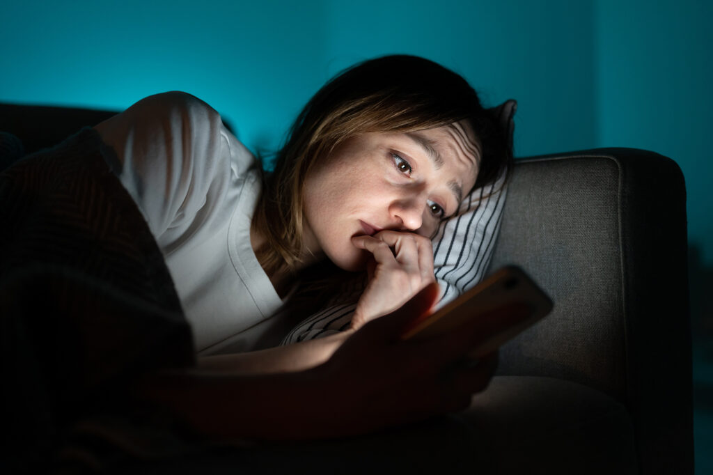 A young woman staring at her phone in the dark while laying on her side in bed. A harsh blue light is emitting from her phone, while a softer blue light is being emitted on the back wall behind her bed.