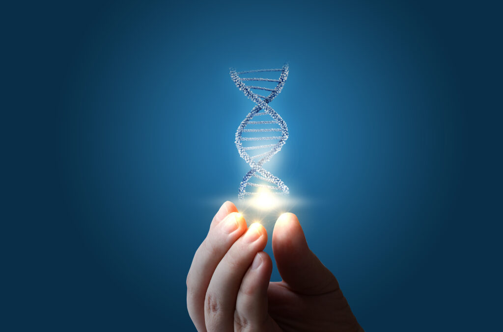 The image shows a close-up of a person’s fingers gently holding a glowing point of light. Above the light, a three-dimensional DNA double helix made of tiny, reflective particles rises upward against a smooth, dark blue background. The scene gives the impression of someone delicately holding or creating genetic material, symbolizing concepts like biotechnology, synthetic biology.