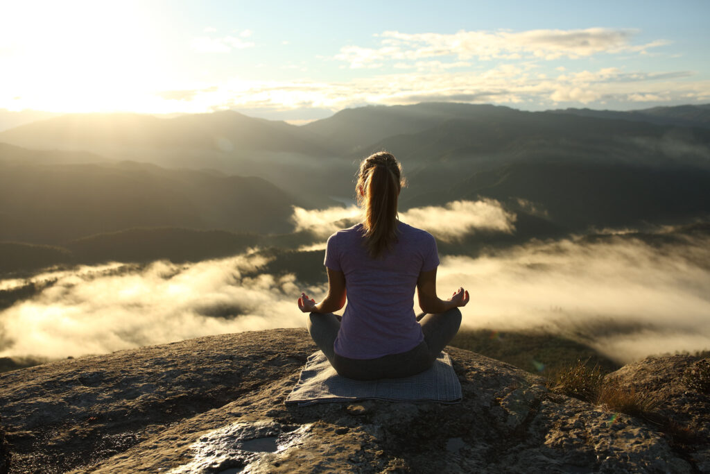 A woman sits cross-legged on a mountain ledge at sunrise, hands resting in quiet meditation. Below her, a sea of clouds drifts through shadowed valleys, while the horizon glows with golden light. The image captures a moment of stillness and balance, symbolizing alignment between body, mind, and the natural rhythm of the day.