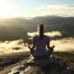 A woman sits cross-legged on a mountain ledge at sunrise, hands resting in quiet meditation. Below her, a sea of clouds drifts through shadowed valleys, while the horizon glows with golden light. The image captures a moment of stillness and balance, symbolizing alignment between body, mind, and the natural rhythm of the day.