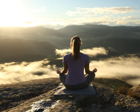 A woman sits cross-legged on a mountain ledge at sunrise, hands resting in quiet meditation. Below her, a sea of clouds drifts through shadowed valleys, while the horizon glows with golden light. The image captures a moment of stillness and balance, symbolizing alignment between body, mind, and the natural rhythm of the day.
