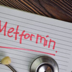 An open spiral notebook rests on a wooden surface, its ruled page filled with the bold red word “Metformin,” underlined for emphasis. A stethoscope, partly visible at the bottom edge, frames the scene and connects the handwritten note to medicine and health. The contrast of red ink against the lined paper suggests urgency and importance, while the rustic grain of the desk background gives the composition a grounded, tactile quality.