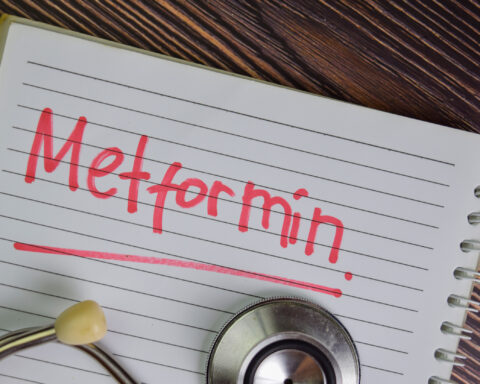 An open spiral notebook rests on a wooden surface, its ruled page filled with the bold red word “Metformin,” underlined for emphasis. A stethoscope, partly visible at the bottom edge, frames the scene and connects the handwritten note to medicine and health. The contrast of red ink against the lined paper suggests urgency and importance, while the rustic grain of the desk background gives the composition a grounded, tactile quality.