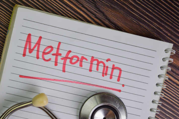 An open spiral notebook rests on a wooden surface, its ruled page filled with the bold red word “Metformin,” underlined for emphasis. A stethoscope, partly visible at the bottom edge, frames the scene and connects the handwritten note to medicine and health. The contrast of red ink against the lined paper suggests urgency and importance, while the rustic grain of the desk background gives the composition a grounded, tactile quality.