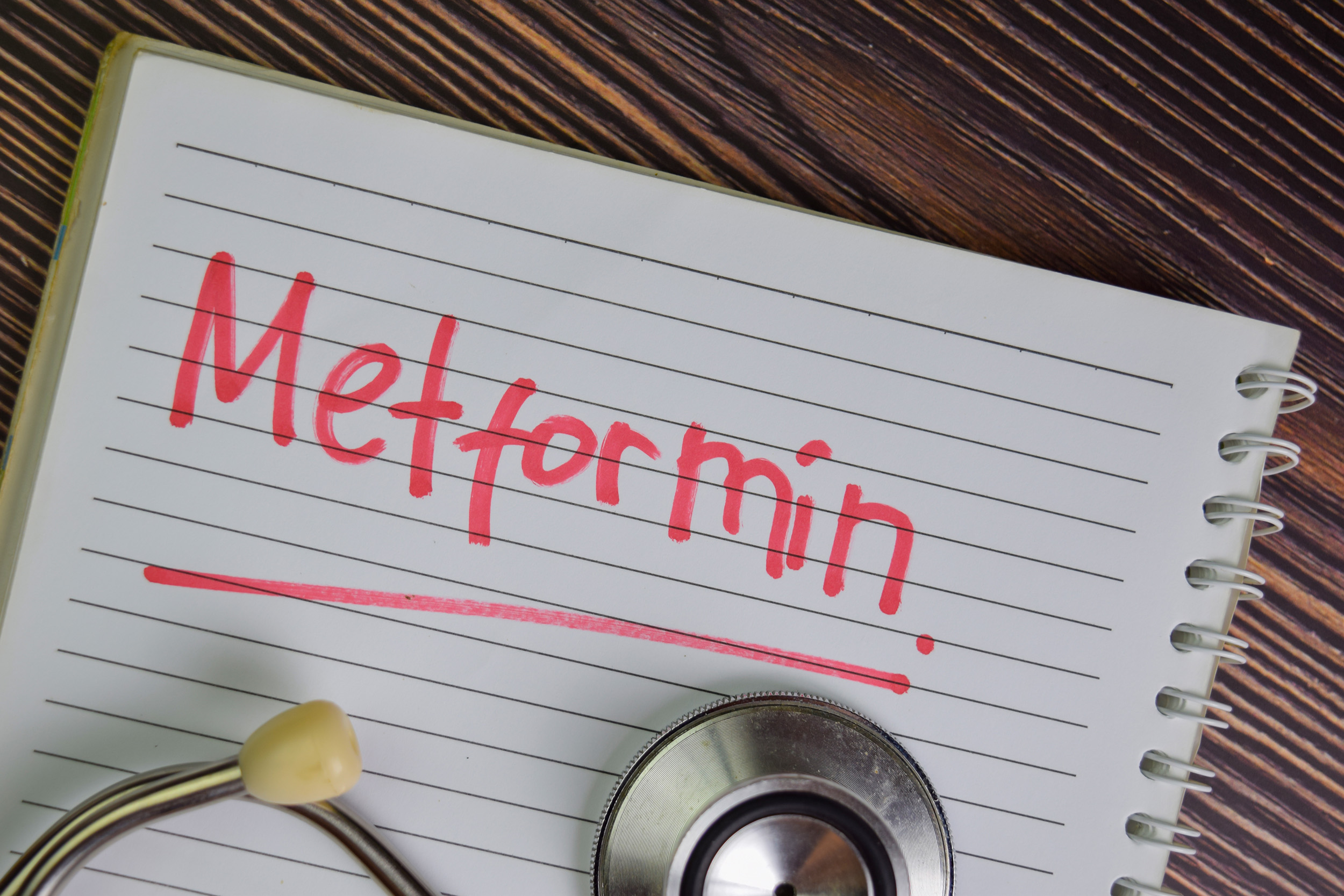 An open spiral notebook rests on a wooden surface, its ruled page filled with the bold red word “Metformin,” underlined for emphasis. A stethoscope, partly visible at the bottom edge, frames the scene and connects the handwritten note to medicine and health. The contrast of red ink against the lined paper suggests urgency and importance, while the rustic grain of the desk background gives the composition a grounded, tactile quality.