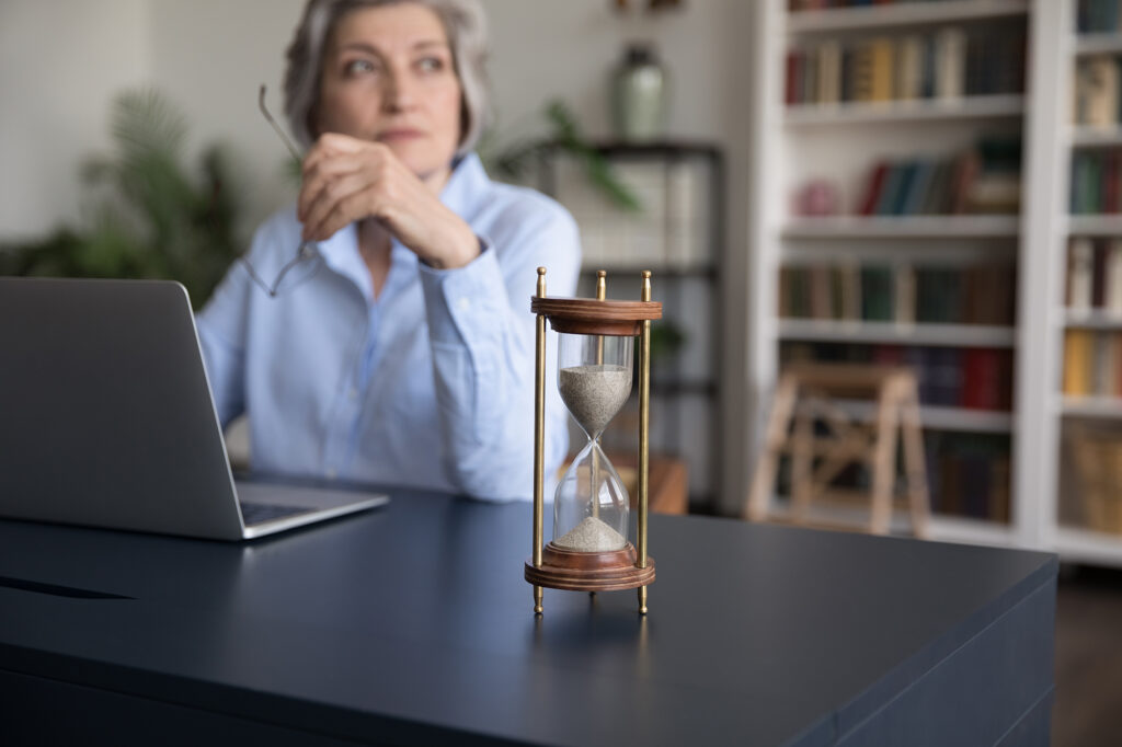 A contemplative older woman sits at her desk, laptop open, glasses in hand, as an hourglass steadily drains on the table before her—a quiet metaphor for time, aging, and the rhythms that govern life.