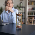 A contemplative older woman sits at her desk, laptop open, glasses in hand, as an hourglass steadily drains on the table before her—a quiet metaphor for time, aging, and the rhythms that govern life.