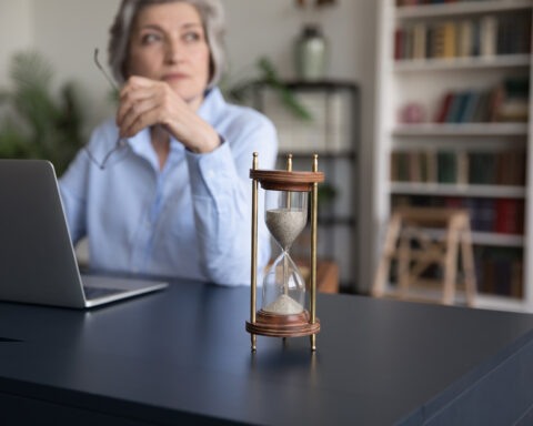 A contemplative older woman sits at her desk, laptop open, glasses in hand, as an hourglass steadily drains on the table before her—a quiet metaphor for time, aging, and the rhythms that govern life.