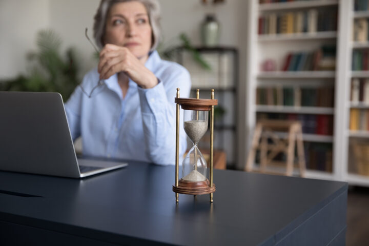 A contemplative older woman sits at her desk, laptop open, glasses in hand, as an hourglass steadily drains on the table before her—a quiet metaphor for time, aging, and the rhythms that govern life.