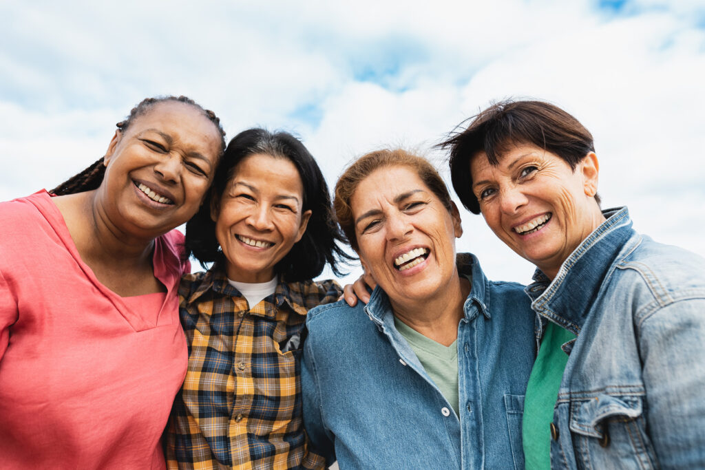 Four women stand shoulder to shoulder beneath a wide blue sky, laughter softening their faces. Their arms rest easily around each other, denim and bright colors mingling, the warmth of friendship and resilience shining through their smiles. The image evokes the vitality and joy of healthy aging.