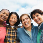 Four women stand shoulder to shoulder beneath a wide blue sky, laughter softening their faces. Their arms rest easily around each other, denim and bright colors mingling, the warmth of friendship and resilience shining through their smiles. The image evokes the vitality and joy of healthy aging.