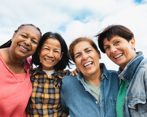 Four women stand shoulder to shoulder beneath a wide blue sky, laughter softening their faces. Their arms rest easily around each other, denim and bright colors mingling, the warmth of friendship and resilience shining through their smiles. The image evokes the vitality and joy of healthy aging.