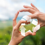 Two hands form a circle against a blurred green landscape, framing a translucent set of recycling arrows that symbolize renewal and continuous cycles of life. Subtle glowing hexagons with icons for health, sustainability, and science float across the foreground, blending natural imagery with futuristic design to evoke the concept of cellular recycling and longevity.