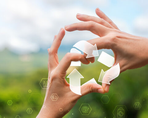 Two hands form a circle against a blurred green landscape, framing a translucent set of recycling arrows that symbolize renewal and continuous cycles of life. Subtle glowing hexagons with icons for health, sustainability, and science float across the foreground, blending natural imagery with futuristic design to evoke the concept of cellular recycling and longevity.