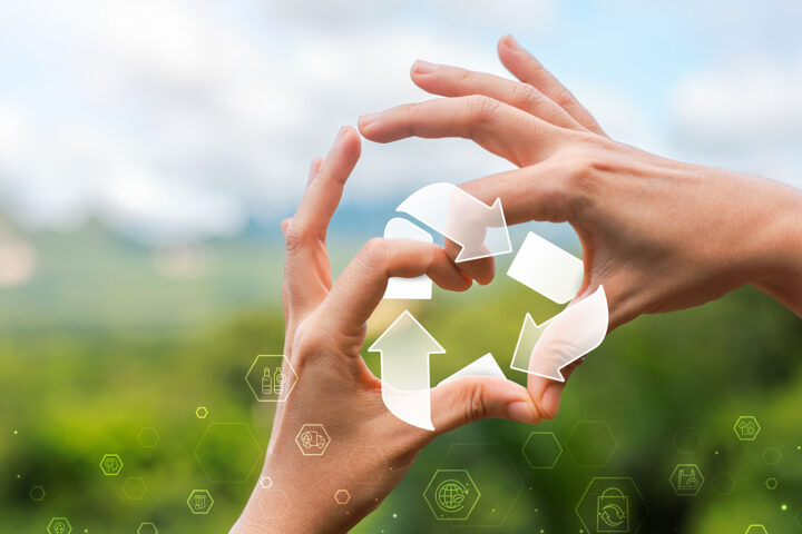 Two hands form a circle against a blurred green landscape, framing a translucent set of recycling arrows that symbolize renewal and continuous cycles of life. Subtle glowing hexagons with icons for health, sustainability, and science float across the foreground, blending natural imagery with futuristic design to evoke the concept of cellular recycling and longevity.